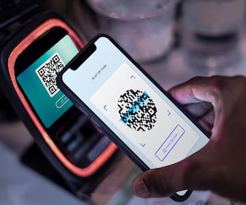 Hand holding smartphone displaying QR code scanner interface next to payment terminal device for contactless mobile payment transaction
