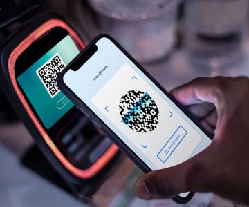 Hand holding smartphone displaying QR code scanner interface next to payment terminal device for contactless mobile payment transaction