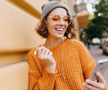 Smiling young woman wearing gray beanie and orange knit sweater, holding smartphone while walking outdoors in urban setting with yellow-tinted sunglasses